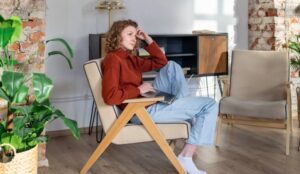 A woman in business casual attire sitting in a tan chair in her living room while holding her laptop in her lap.