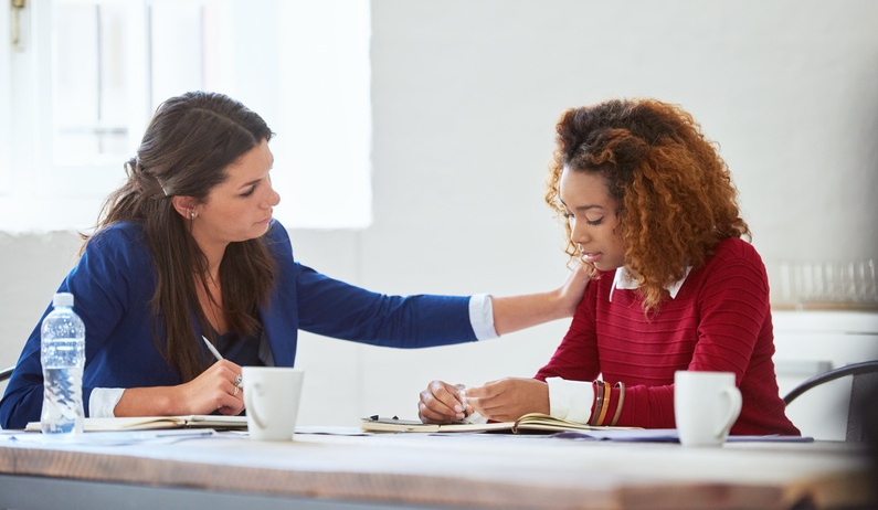 Two professional women sit next to each other at a table. One woman places her hand on the other's shoulder.