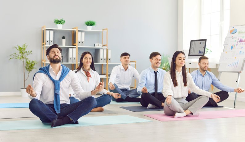 Adults in professional clothing sitting cross-legged on colorful yoga mats inside a large, well-lit office.