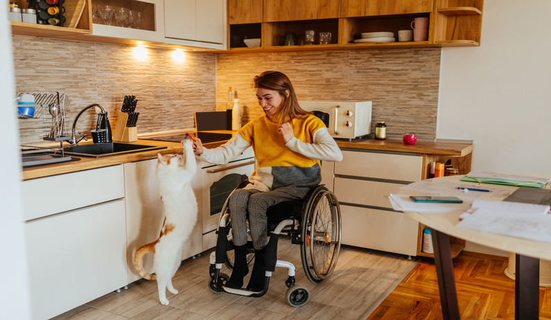 A smiling woman wearing a yellow sweater sits in a wheelchair in a kitchen, playing with an orange and white cat.