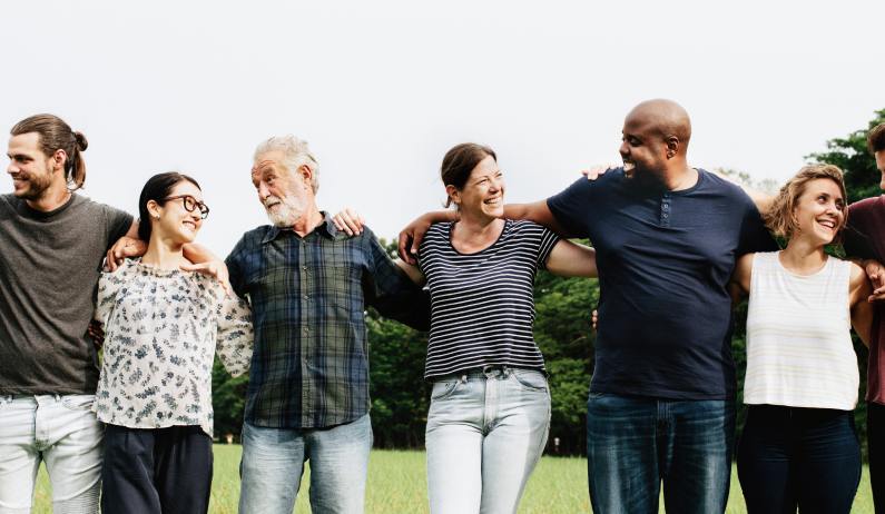 Eight happy people stand in a line with their arms wrapped around the next person's shoulders. They're standing outside.