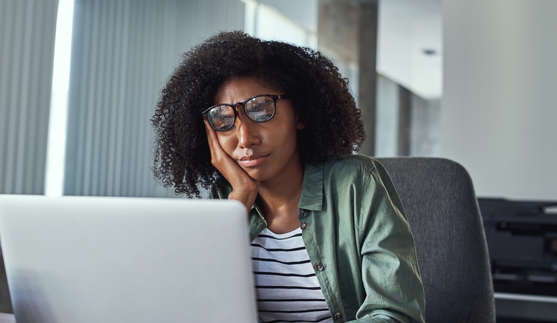 A frustrated worker sitting in front of an open laptop. They are resting their face in their open palm.