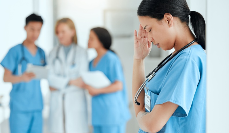 A woman working in healthcare holds her hand against her head. She leans against a wall in front of other workers.