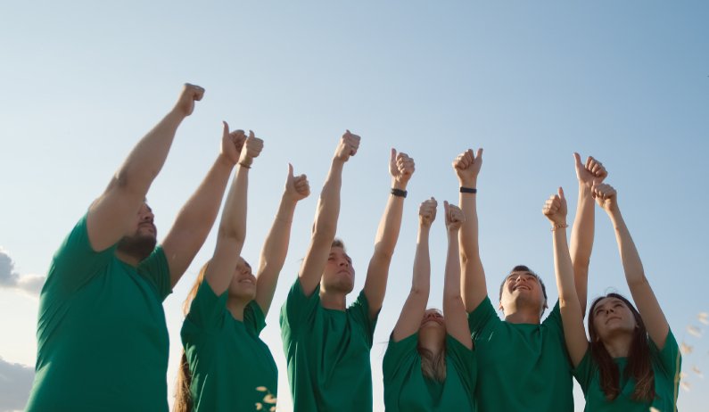 A group of smiling young men and women stand outside in green shirts with arms raised, giving thumbs up.
