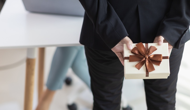 A business person in a sleek black suit holding a ribbon-wrapped present behind their back while standing in a coworker's office.