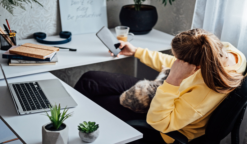 A remote worker sitting in their workspace and looking frustrated as they check their smartphone.