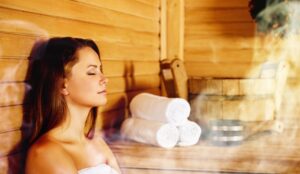 A young, Caucasian woman with brunette hair sits in a sauna with a white towel on and her eyes closed.