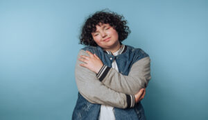 A woman with black curly hair hugging herself. She is wearing a jean jacket and standing in front of a blue wall.