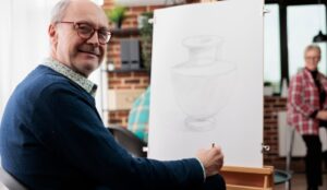 An older man wearing glasses and smiling with a canvas on a stand in front of him while attending a painting class.