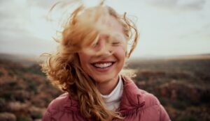 A person smiles with their eyes closed outside. They're wearing a puffer jacket, and the wind is blowing their hair in their face.