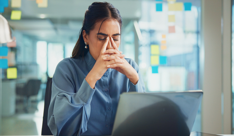 A person sitting in a tidy office space and looking stressed by a laptop, with their fingers against the bridge of their nose.