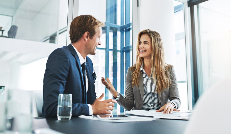 A woman wearing a blazer sits at a desk and speaks to a man wearing a navy suit. Papers and a tablet are on the table.