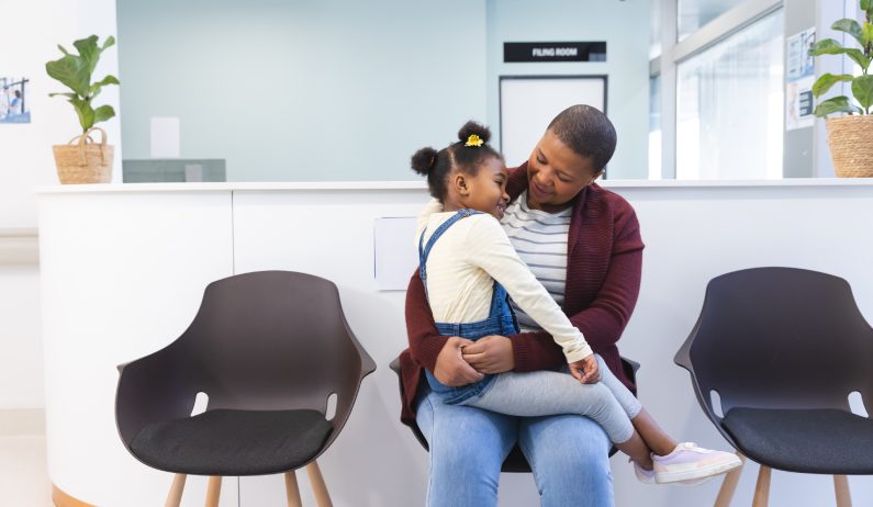 A parent sitting in a medical waiting room holding a young child on her lap, both smiling and appearing calm.