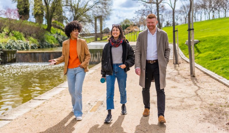 Three adults walking along park path by water, talking and smiling, with trees, grass, and a bridge in the background.