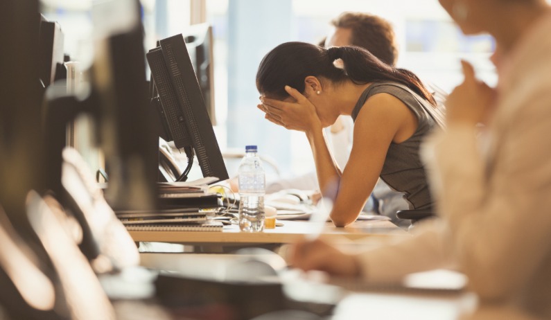 A woman in a shared office space leans forward with her elbows on her desk and her face in her hands.