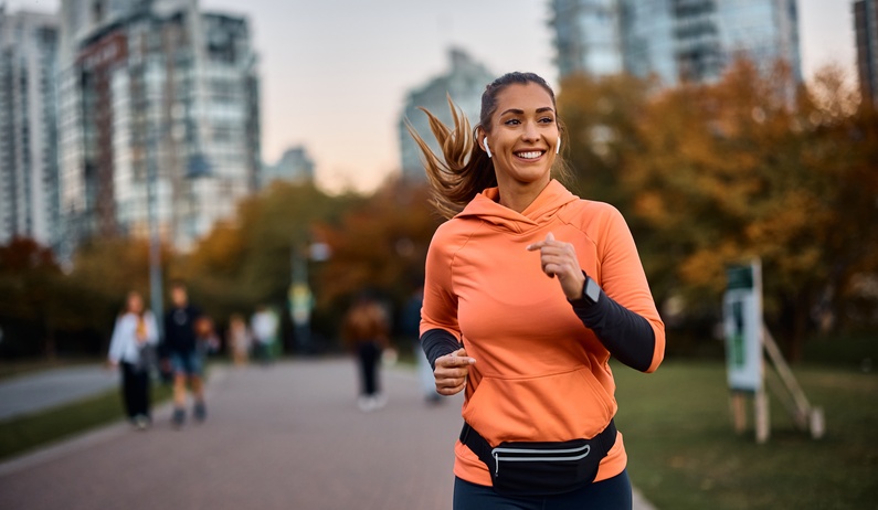 A woman runs along a paved path in a park, wearing earbuds and athletic clothing, with trees and buildings in the background.