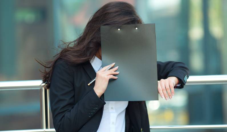 A professional person standing against a railing in an office and hiding their face with a clipboard or binder.