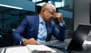 A business professional sits at a laptop with eyes closed, appearing stressed during a late office work session.