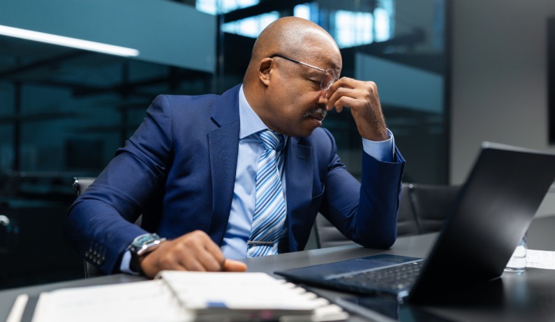 A business professional sits at a laptop with eyes closed, appearing stressed during a late office work session.