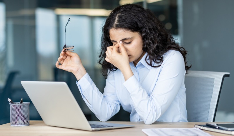 A woman sitting at an office desk, holding glasses and rubbing her eyes beside a laptop, papers, and a pen holder.