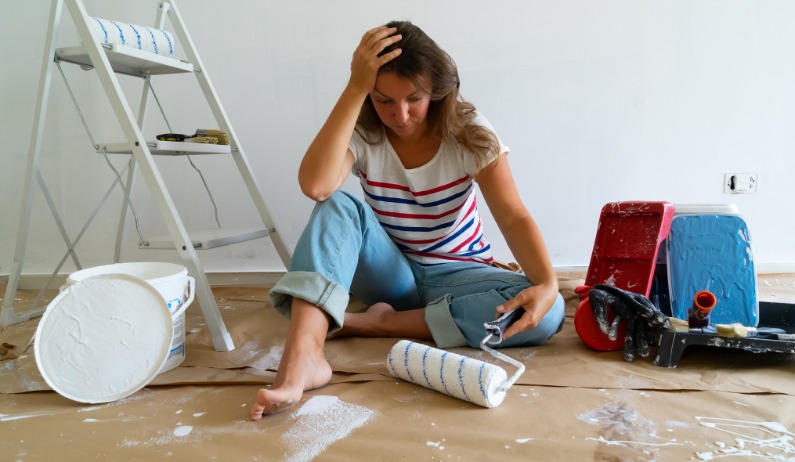 a woman sitting on a covered floor beside paint rollers, buckets, trays, and a ladder in a white room.
