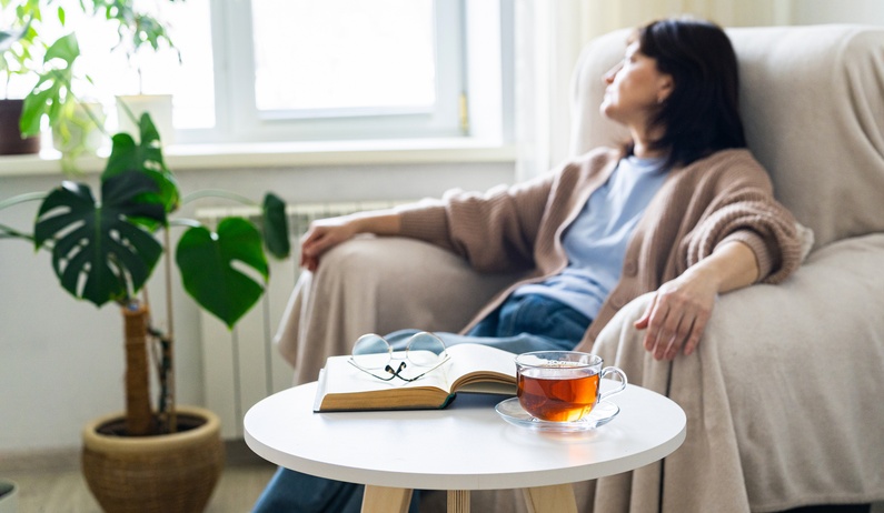 A woman sitting on a couch covered with a blanket, looking at the window next to a coffee table with a book and tea.