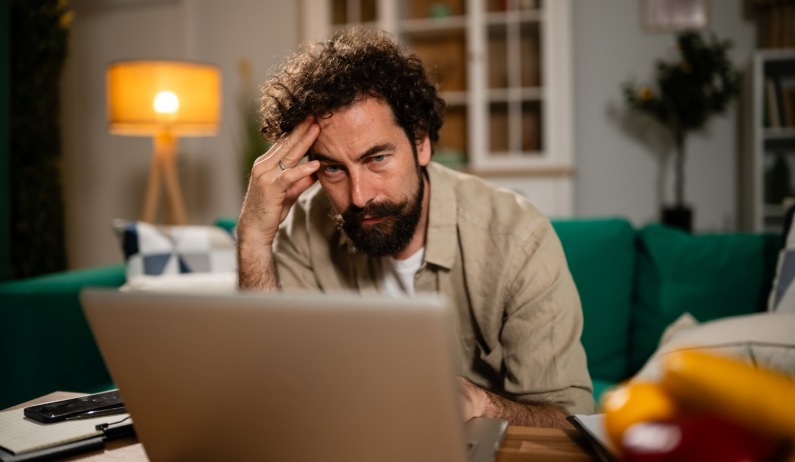 A man in a living room sits on a sofa and uses a laptop on a coffee table. He presses his fingers to his temple.
