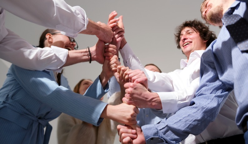 A group of people in dress shirts standing in a circle and stacking hands together against a plain gray background.
