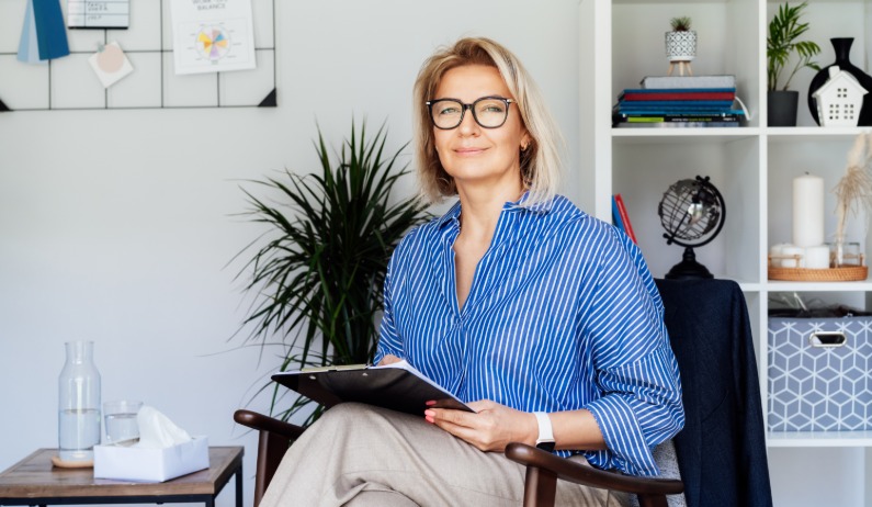 A psychiatrist wearing glasses and a watch holds a notepad in an office. The psychiatrist sits in a chair.