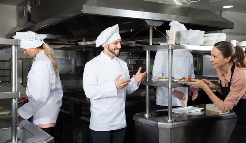 A man in a white chef's coat gesturing toward a woman holding a plate of food in a kitchen with other workers.